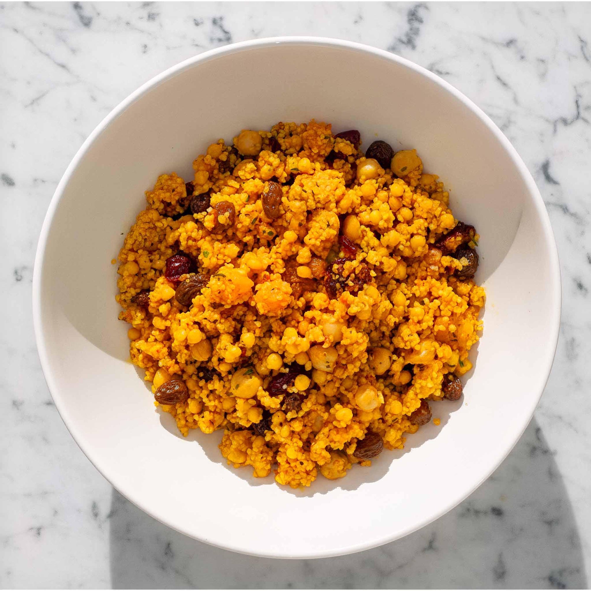 White bowl filled with spiced couscous with chickpeas and raisins on a marble surface.