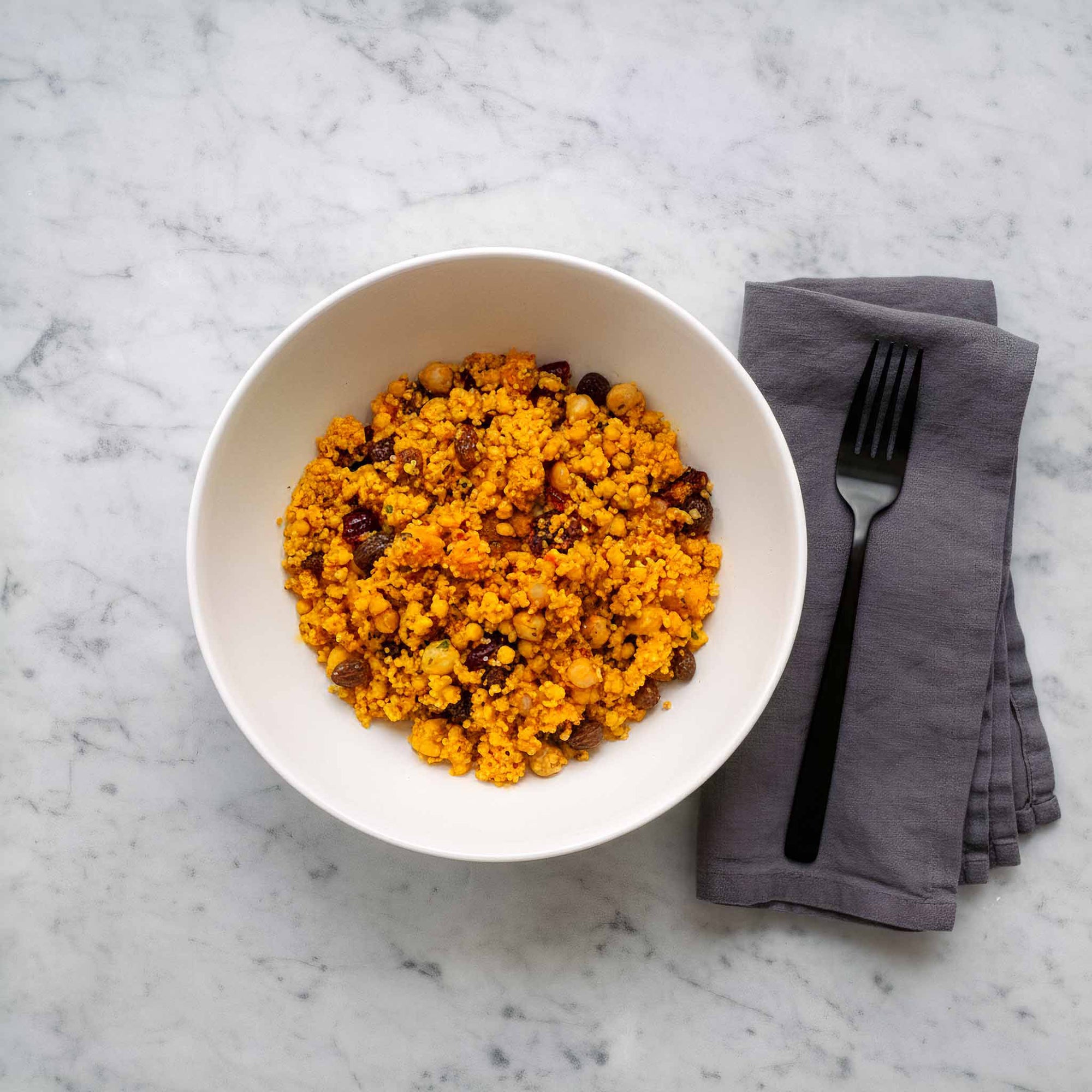 White bowl with spiced couscous with chickpeas and raisins on a marble surface with a gray napkin and fork.