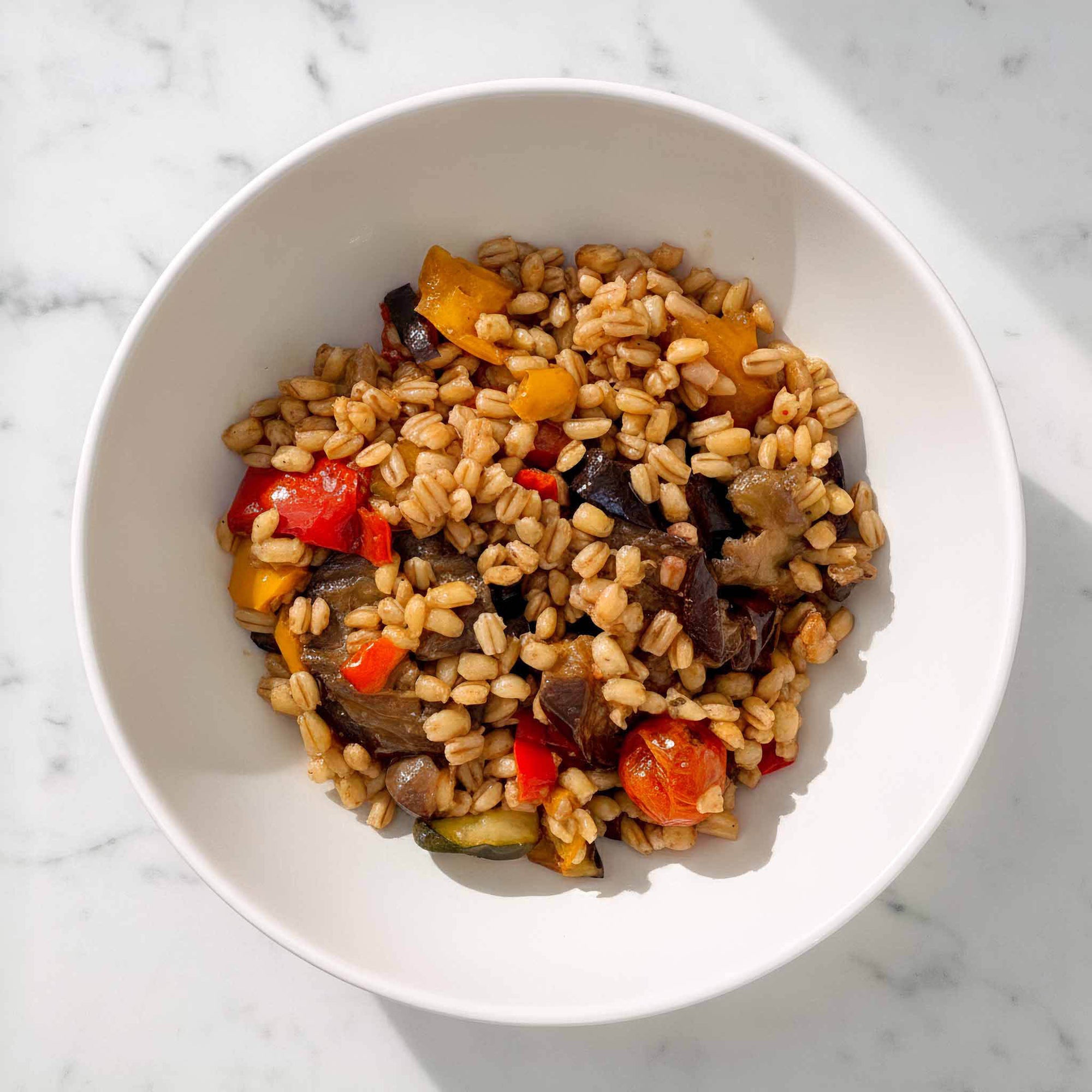 Close-up of barley salad with roasted vegetables, balsamic glaze, and herbs in a white bowl, on a marble surface.