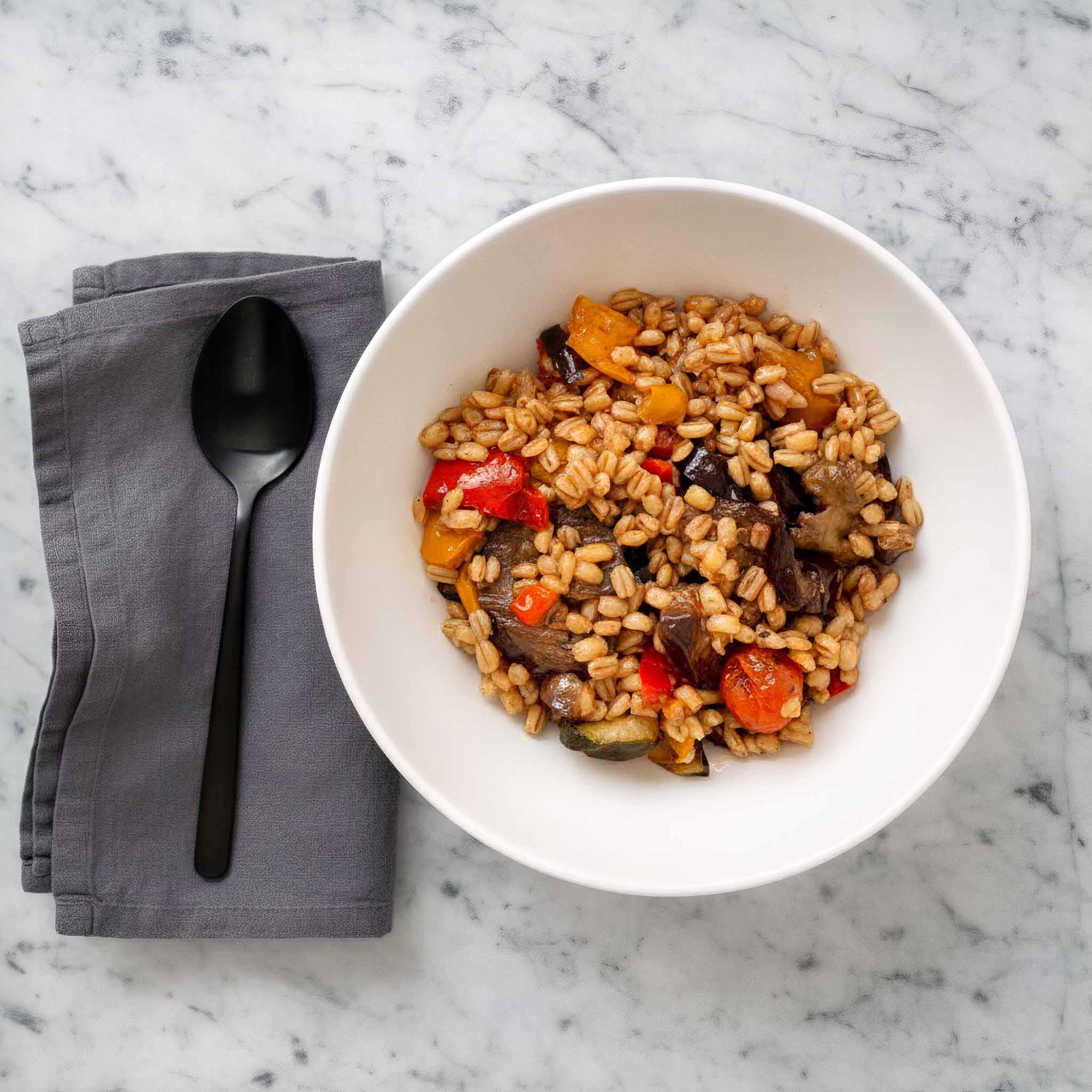 Airheads barley Mediterranean vegetable salad with balsamic glaze in a ceramic bowl on a marble surface with a gray napkin and spoon.