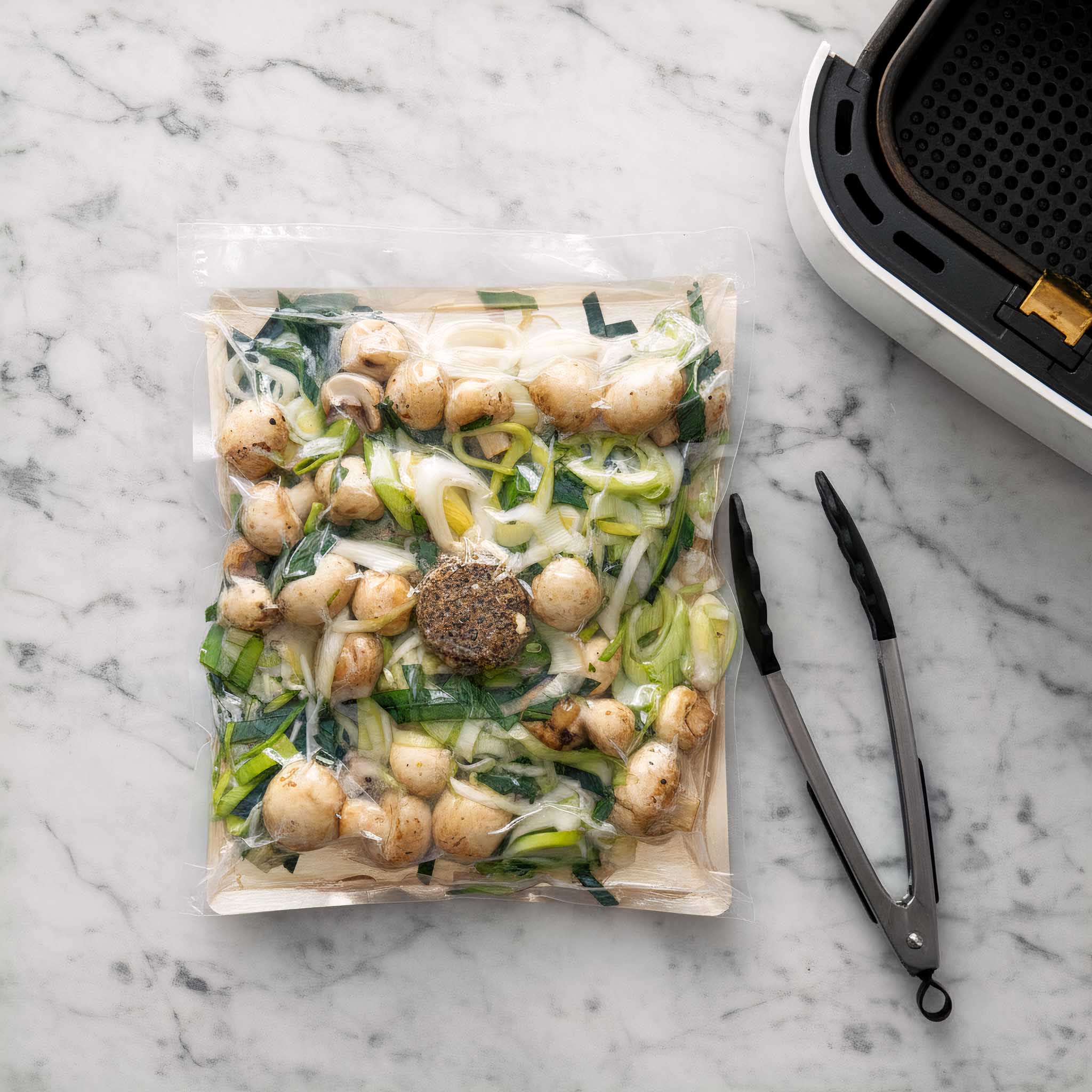 Packaged vacuum-sealed Airheads buttered leaks and mushrooms on a marble surface with tongs and an air fryer.