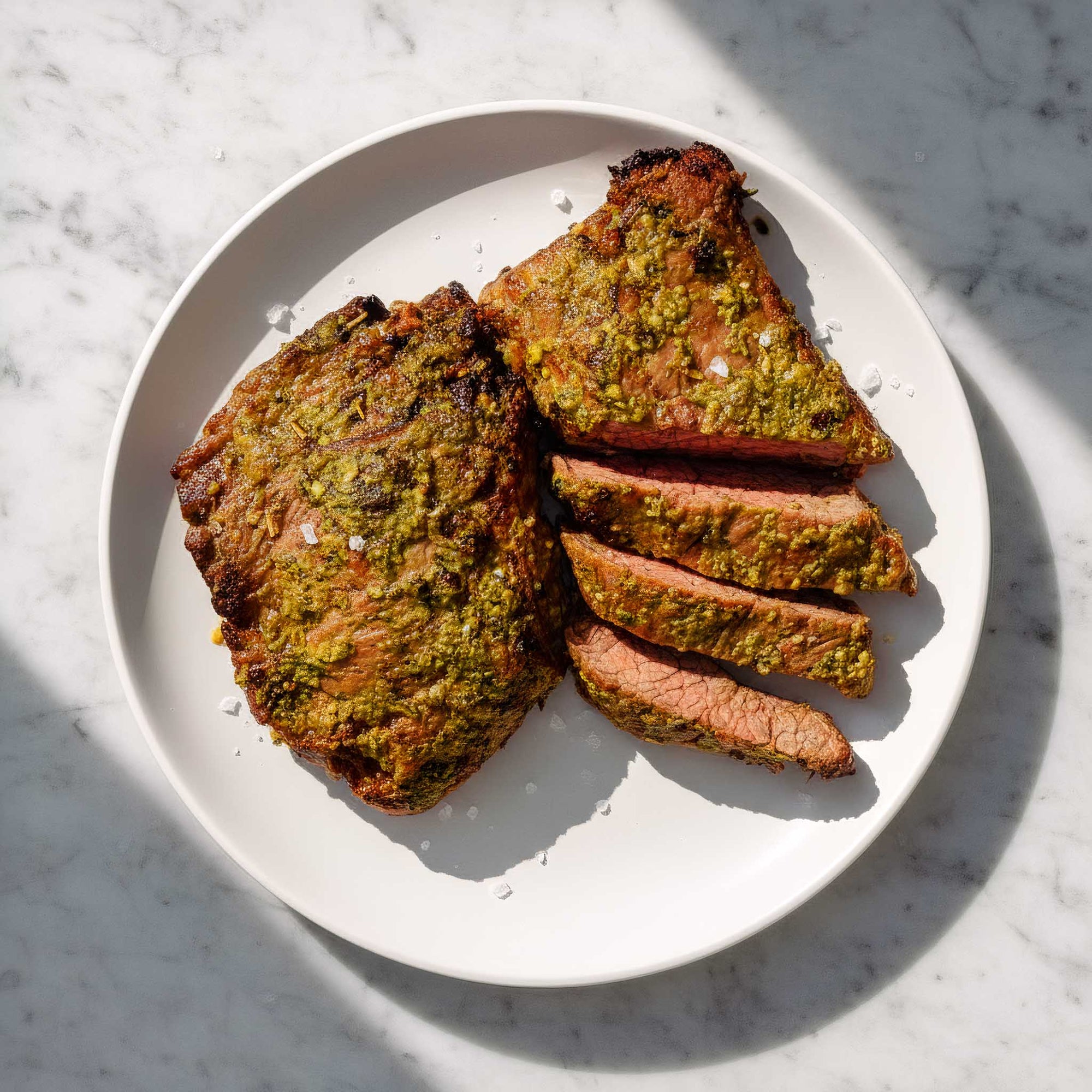 Close-up of chimichurri flank steak with green herb crust and juicy sliced interior on a marble surface. 