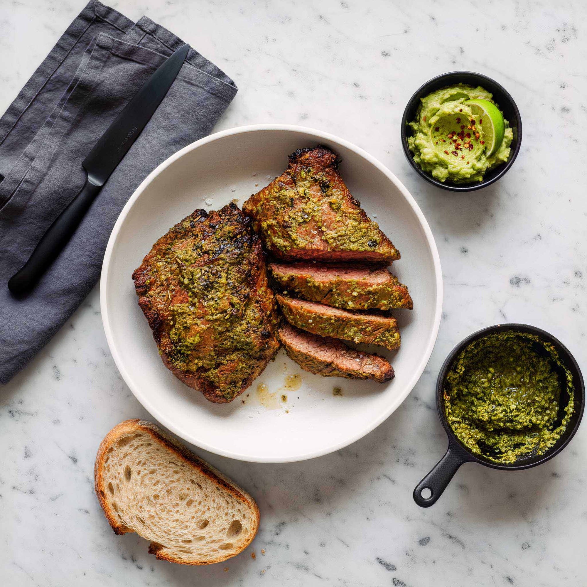 Plated dish of Airheads chimichurri flank steak served with herb sauce, smashed avocado and bread on a marble surface.