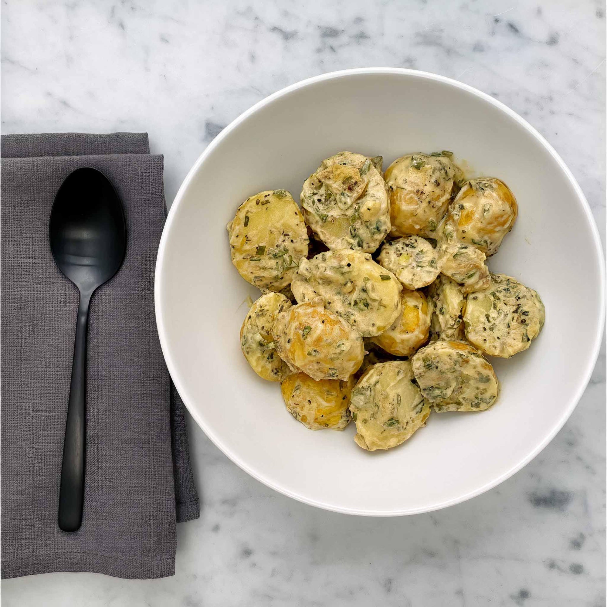 White bowl with dijon mustard potato salad on a marble surface with a black spoon and napkin.