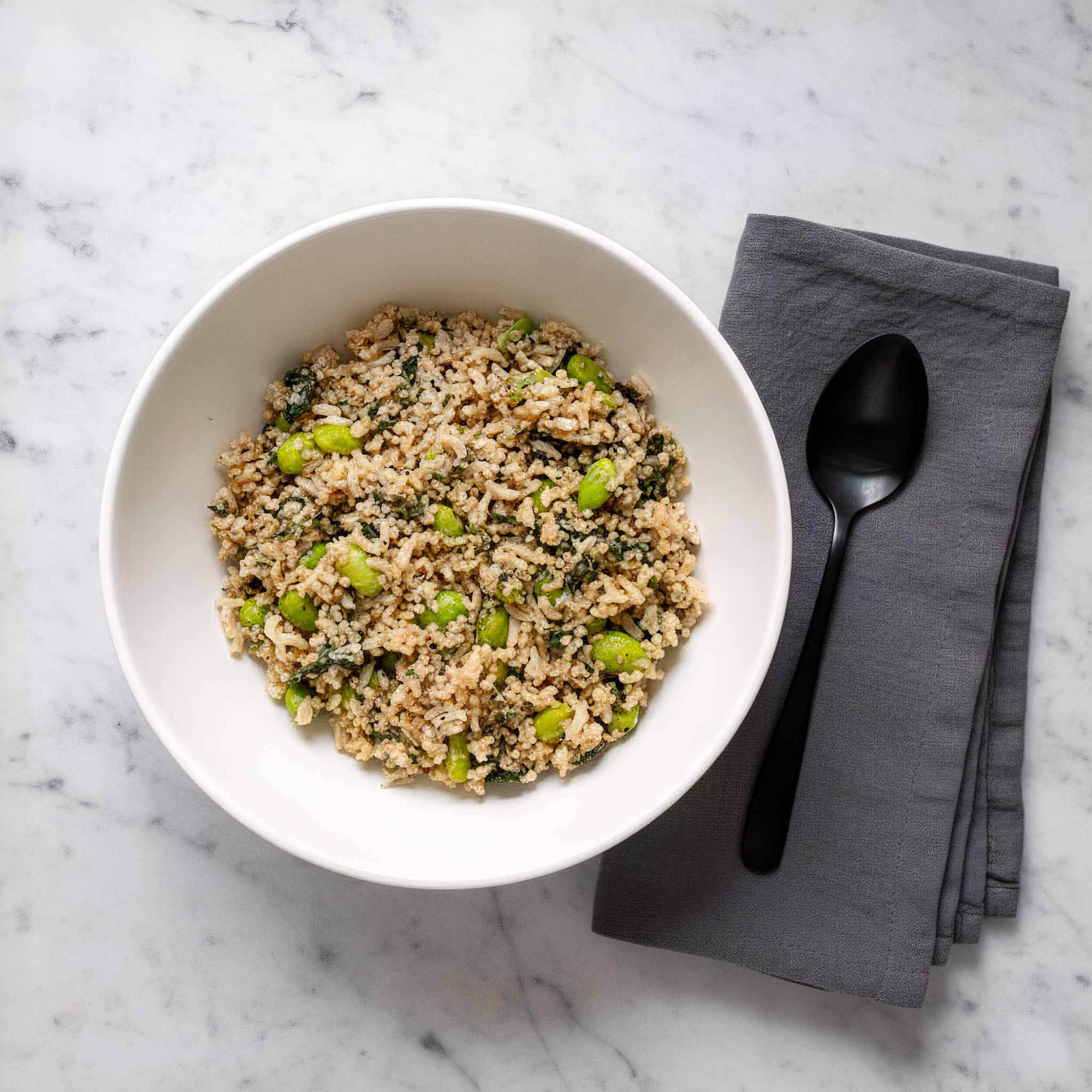 White bowl with edamame quinoa and brown rice salad in a white ceramic bowl on a marble surface with a black spoon and napkin.