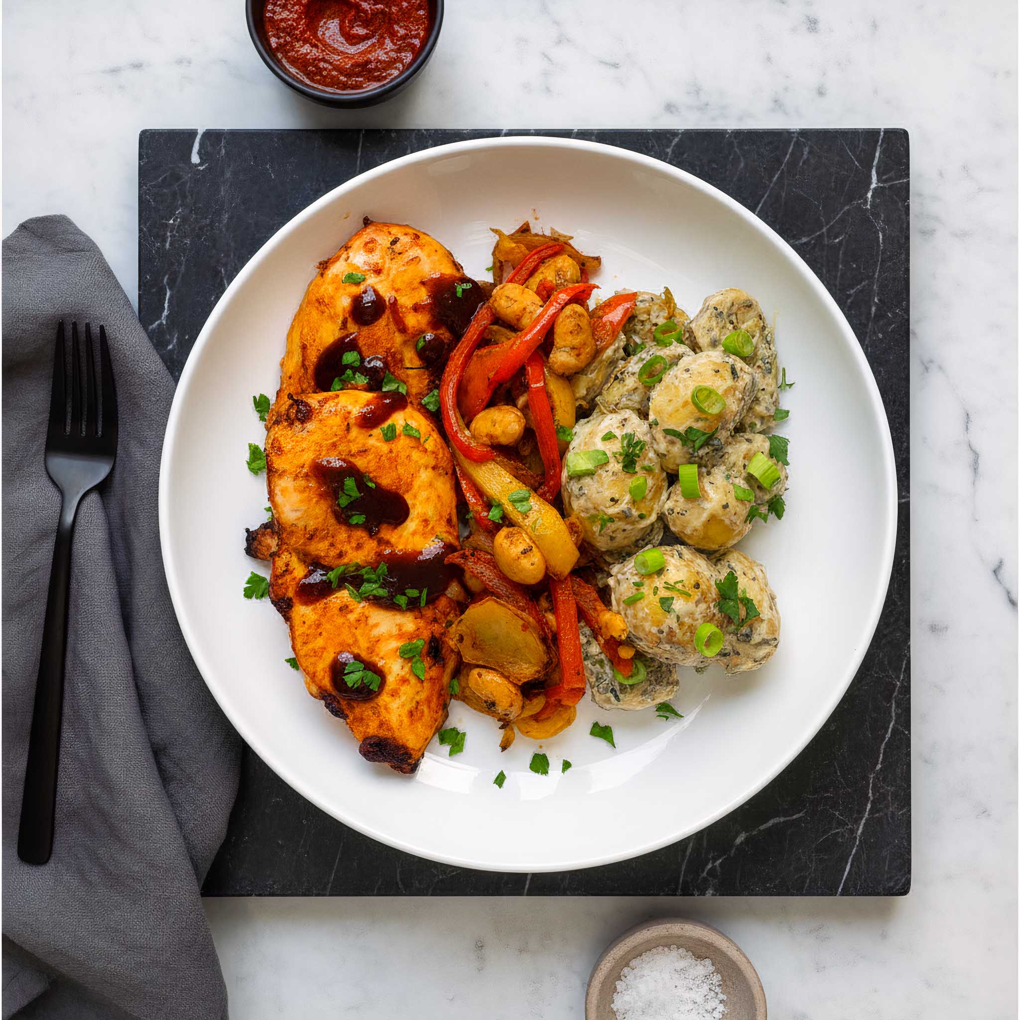 White plate of harissa marinated chicken breasts, Tex-Mex fajita sliced peppers and onions, and dijon potato salad, on a black tray on a marble surface.