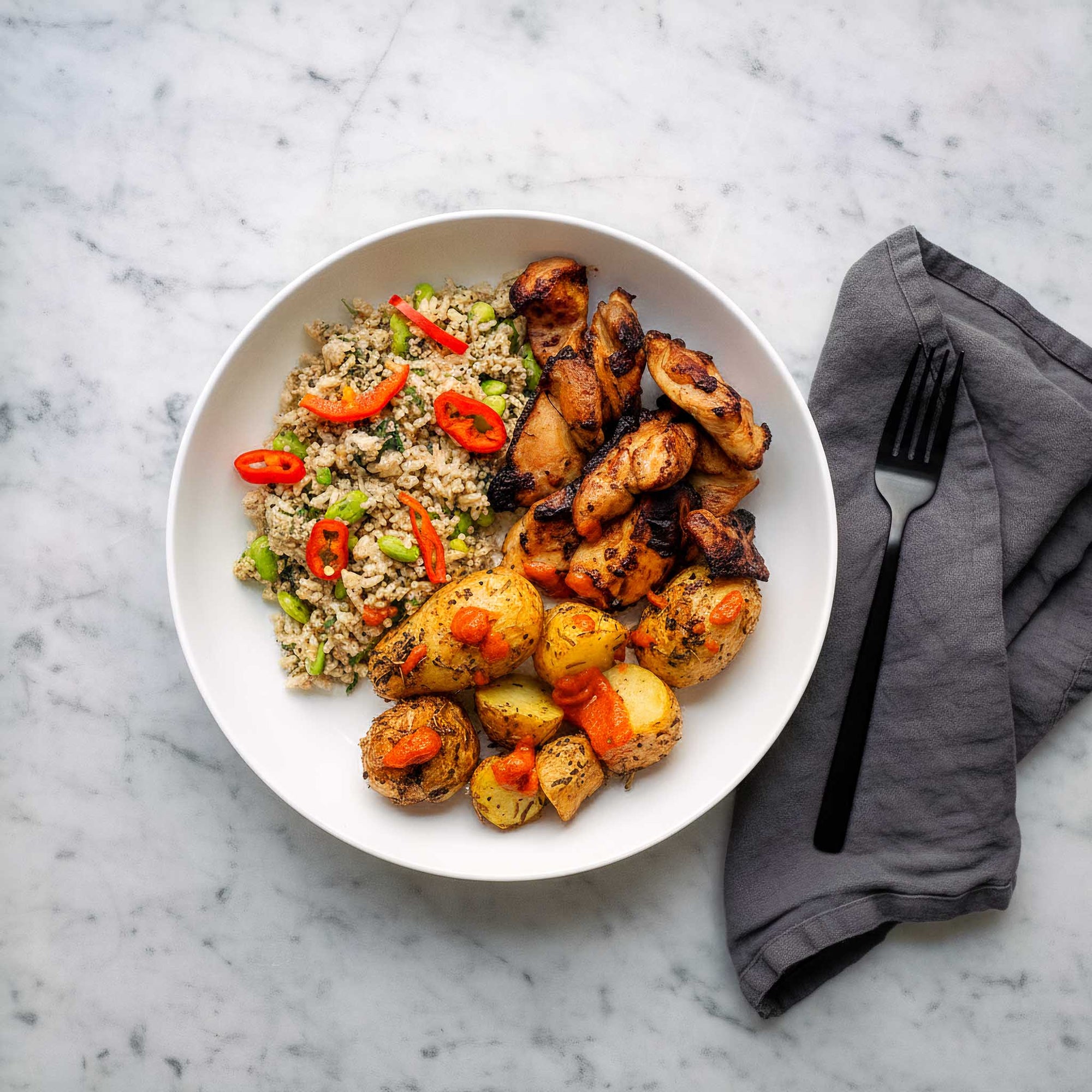 Honey mustard chicken thighs with lemon roast potatoes and green grain salad served in a white bowl on a marble surface.