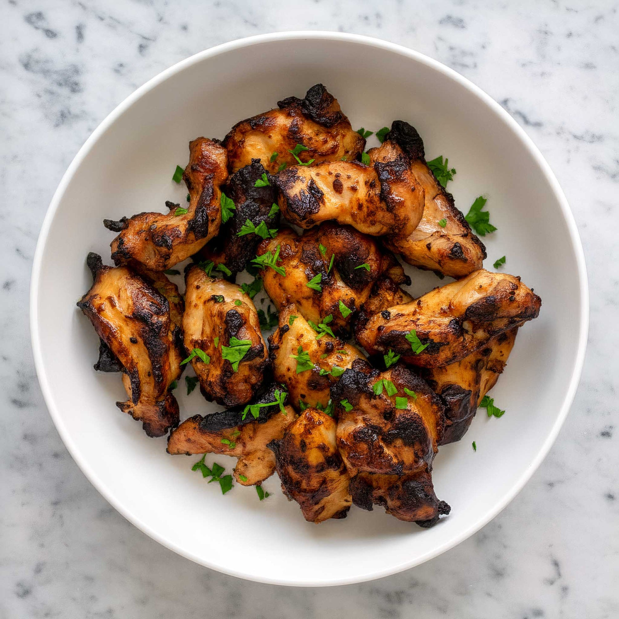 Grilled honey mustard glazed chicken thighs with herbs in a white bowl on a marble background.
