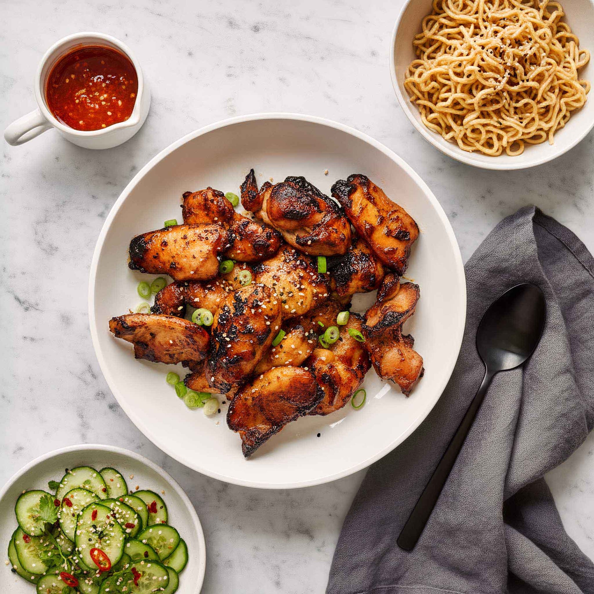 Plated dish of honey sesame chicken thighs with garnishes on a marble surface, accompanied by noodles and a cucumber salad.