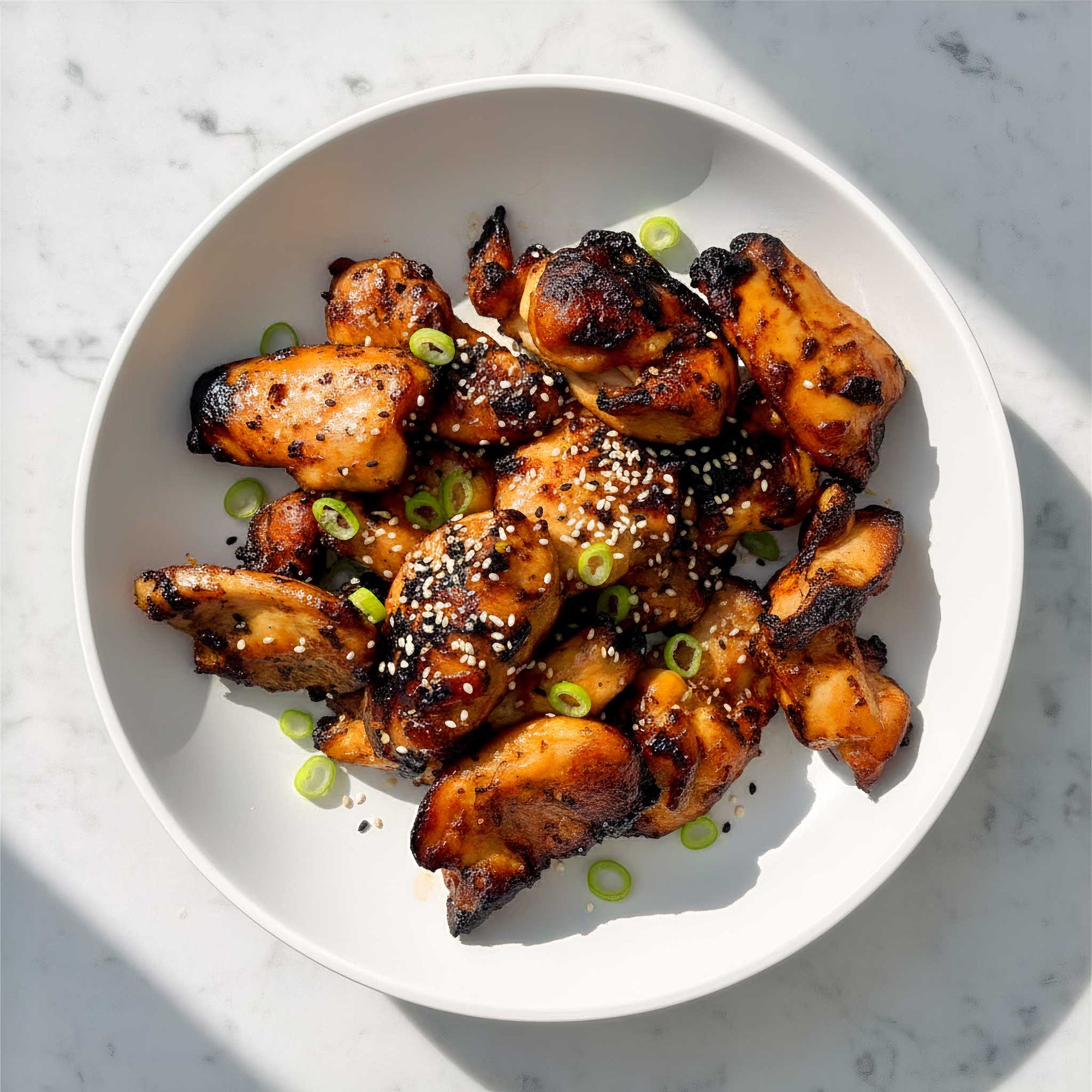 White bowl filled with air fried honey sesame chicken thighs with toasted seeds garnished with green spring onions on a marble surface.