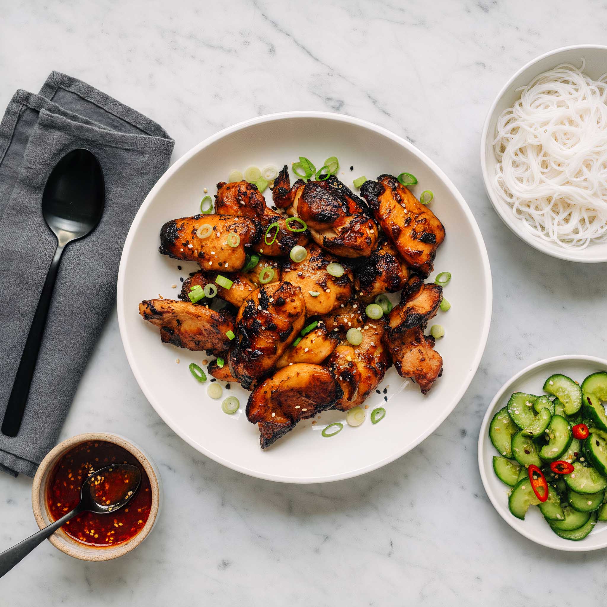 Plated dish of honey sesame chicken thighs with garnishes on a marble surface, accompanied by noodles and a cucumber salad.
