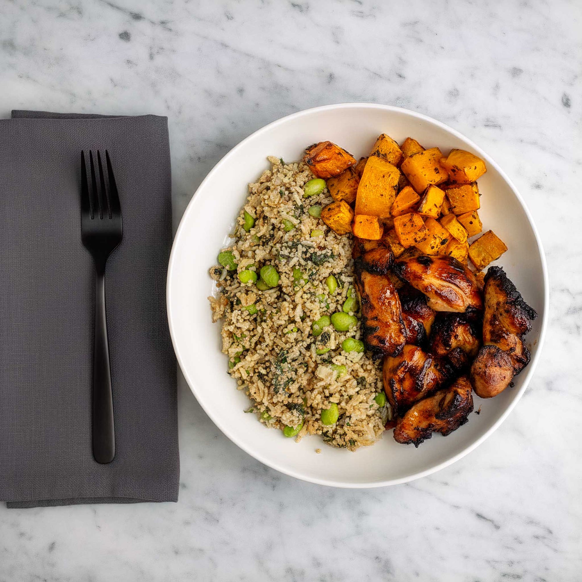 White bowl of chilli honey marinated air fried chicken thighs, garlic and herb carrots and butternut squash, with brown rice and quinoa edamame salad, on a black try on a marble surface.