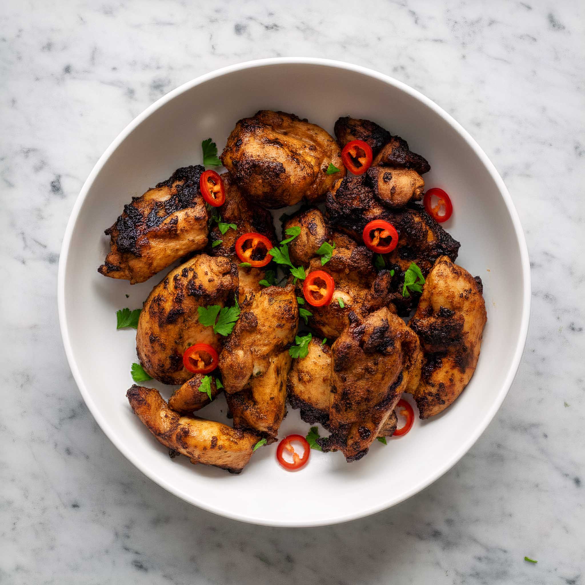 Close-up of jerk chicken thighs with chillis and chopped parsley in a white bowl on a marble background.