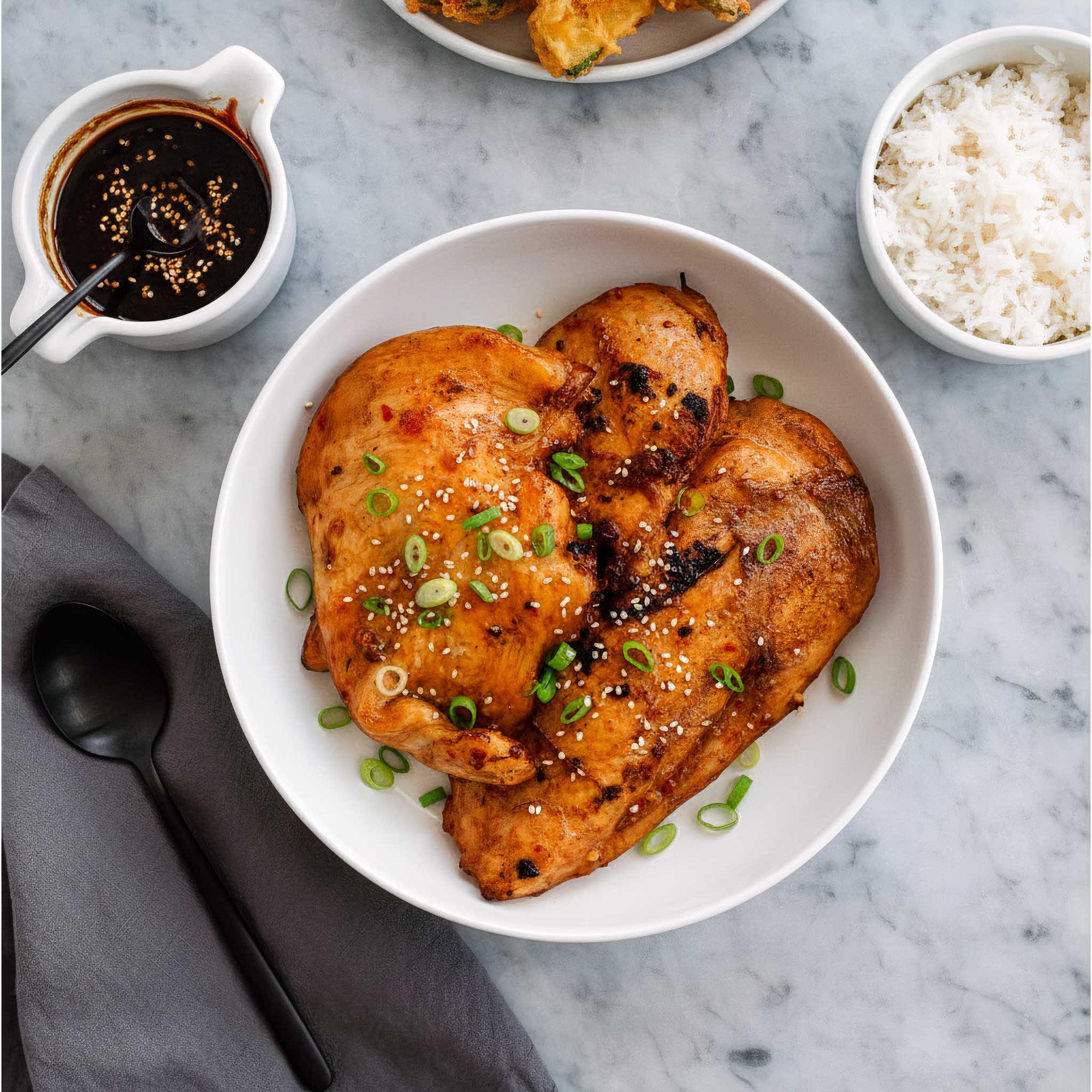 Bowl of Korean BBQ marinated chicken breast with garnishes on a marble surface, accompanied by a small bowl of sauce and rice.