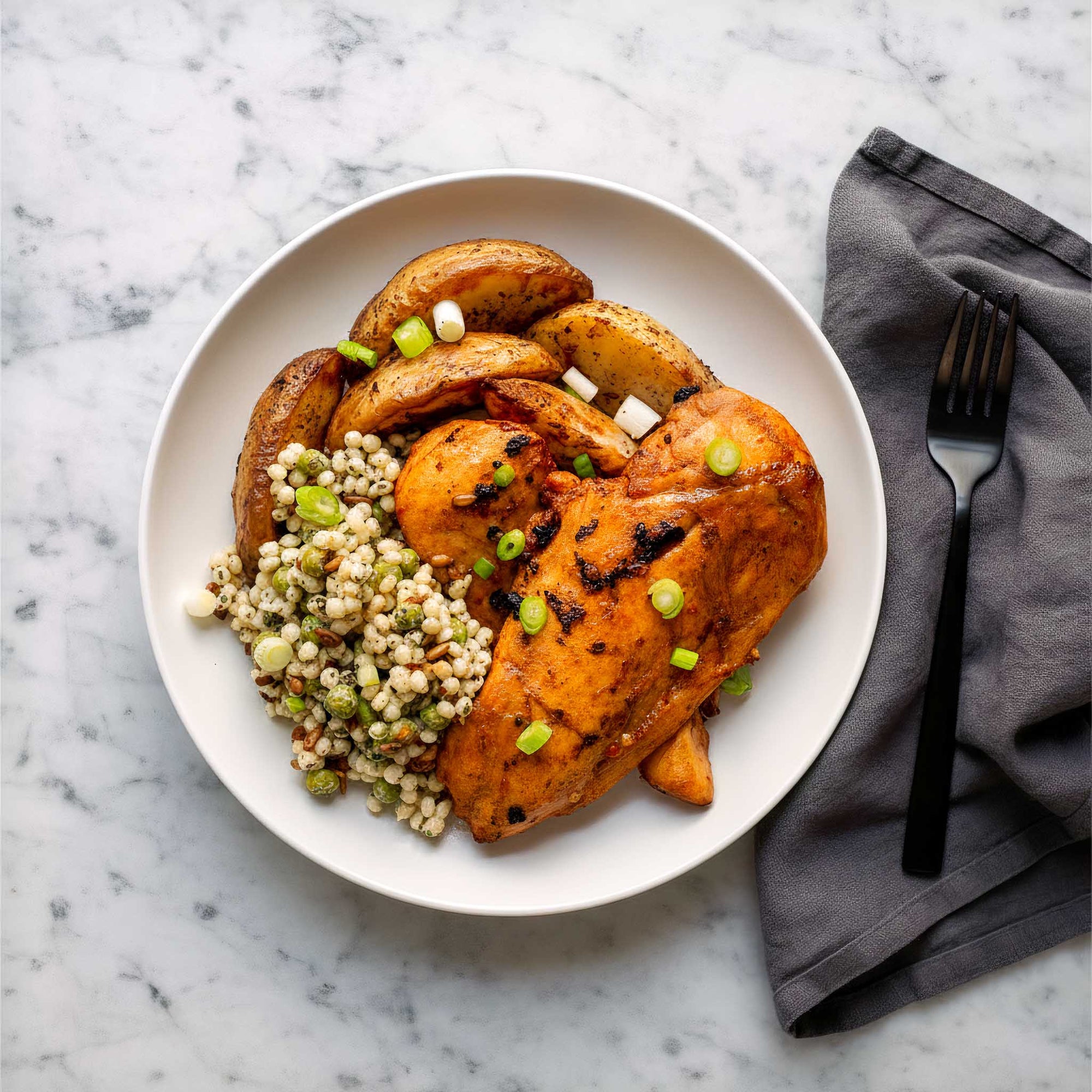 Plated dish with air fried Korean BBQ chicken breasts, sumac roasted potato wedges, and giant minted couscous salad on a marble surface.