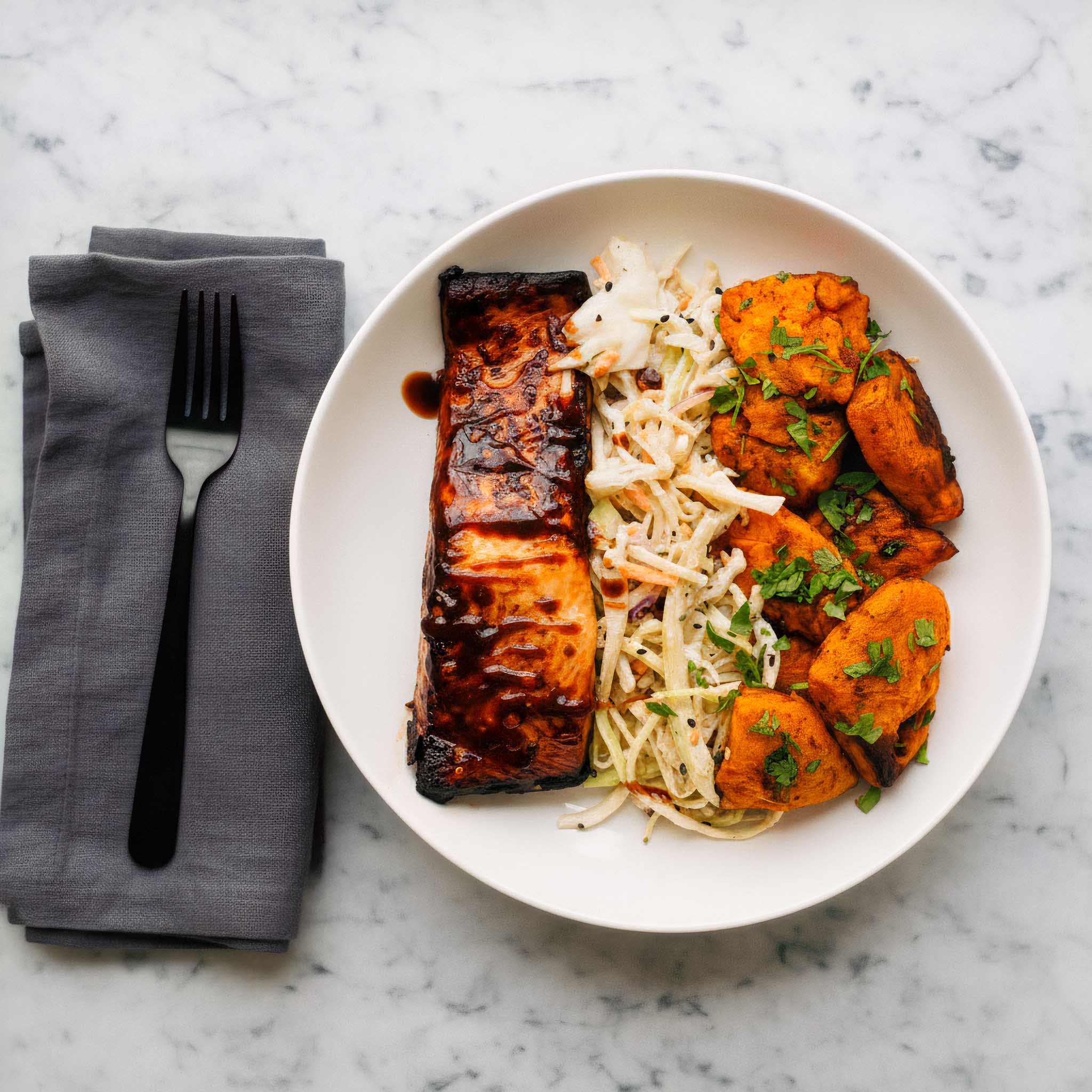White plate with air fried sticky teriyaki salmon fillets, hot shake sweet potatoes and maple mustard coleslaw, on a marble surface with a gray napkin and fork.
