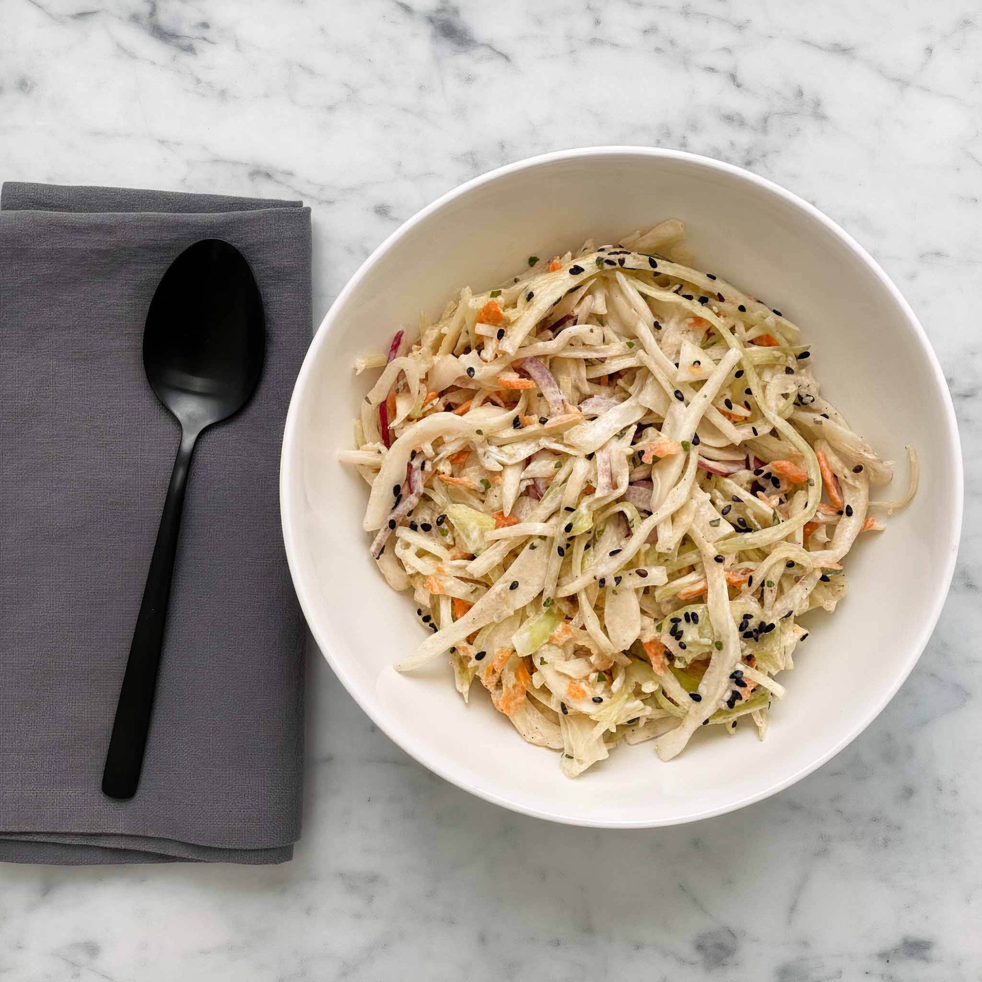 White bowl of maple mustard coleslaw with a black spoon and gray napkin on a marble surface.