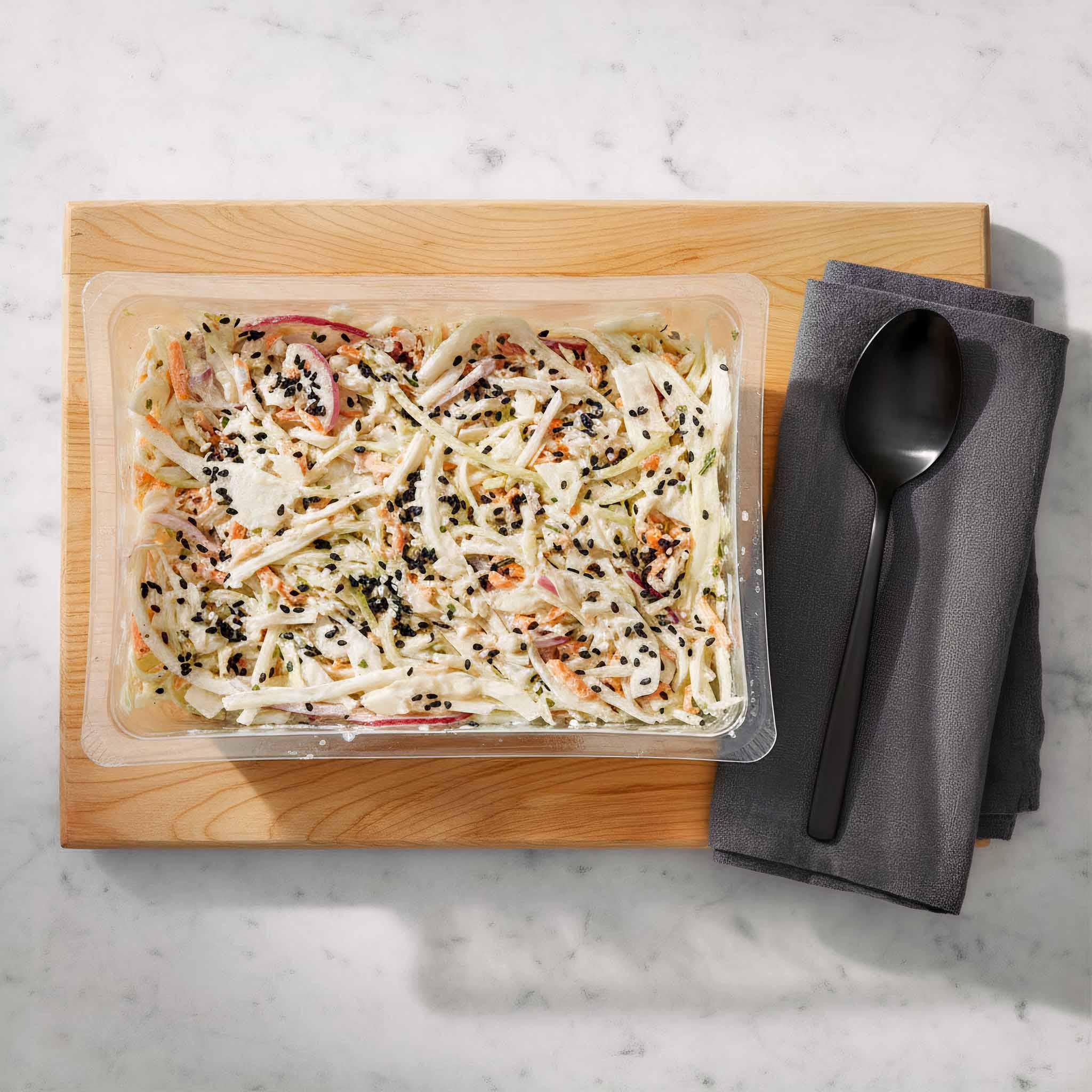 Prepared maple mustard coleslaw in a clear container on a wooden cutting board with a black spoon and napkin.