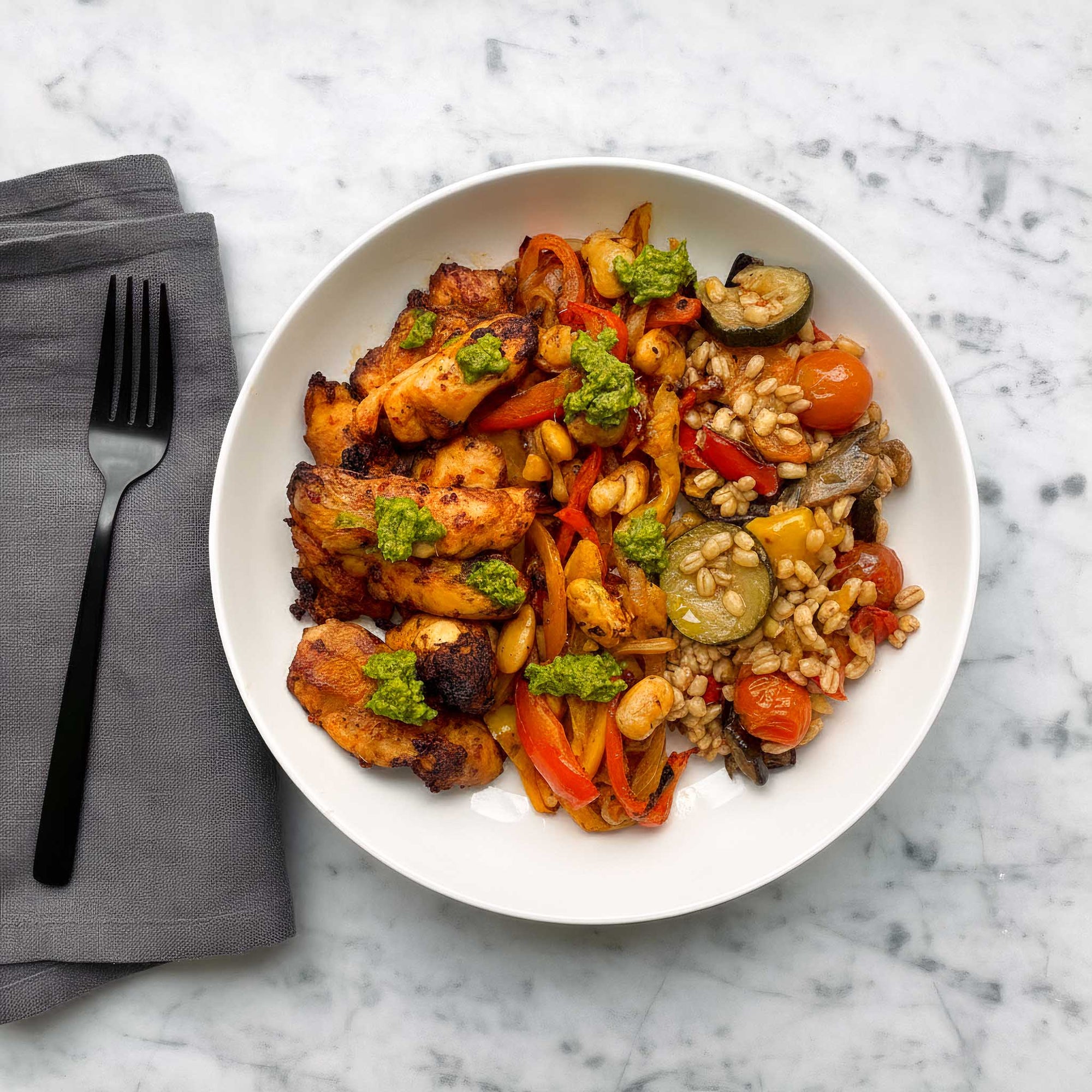 White bowl filled with air fried peri peri chicken thighs, roasted Mediterranean vegetable and barley salad, and fajita spiced peppers and onions on a marble surface with a black fork and grey napkin.