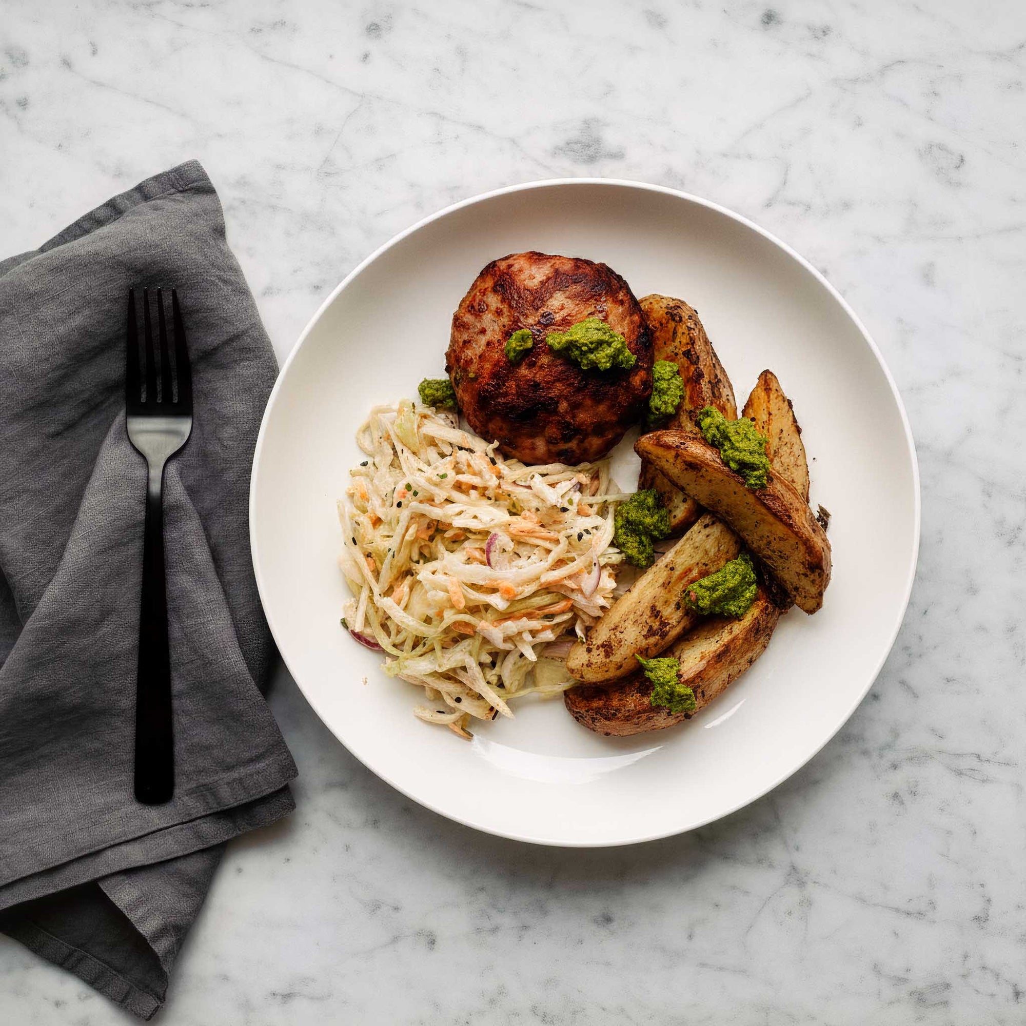 White plate of food with smokey pork and bacon patty, maple mustard coleslaw, and sumac roasted potato wedges on a marble surface, topped with green sauce.