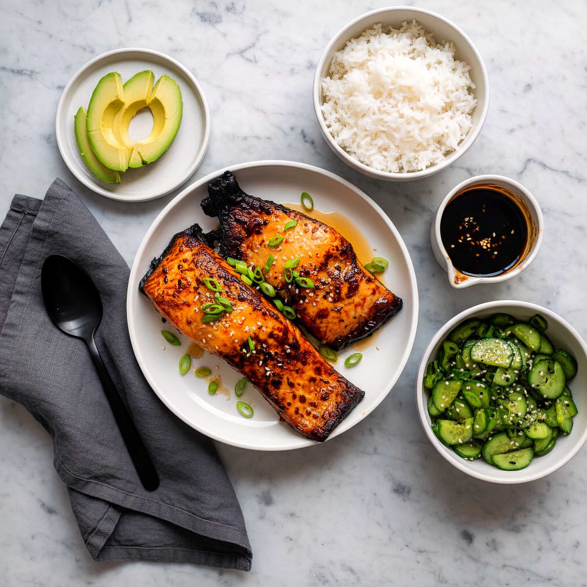 Plated sticky teriyaki salmon fillets with sides of rice, avocado, and cucumber salad on a marble surface.