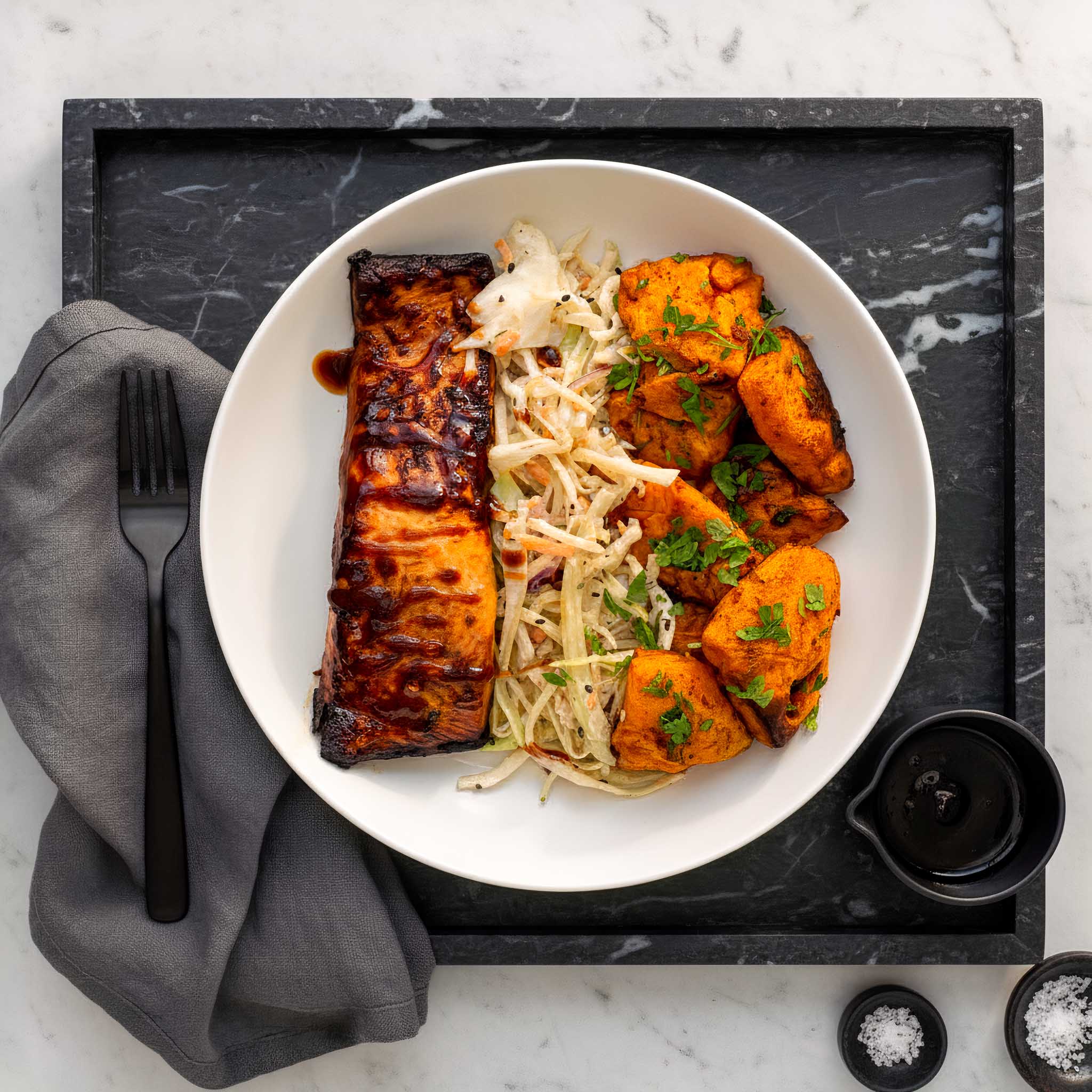 White plate of teriyaki salmon fillets, hot shake sweet potatoes and maple mustard coleslaw, on a black tray on a marble surface.