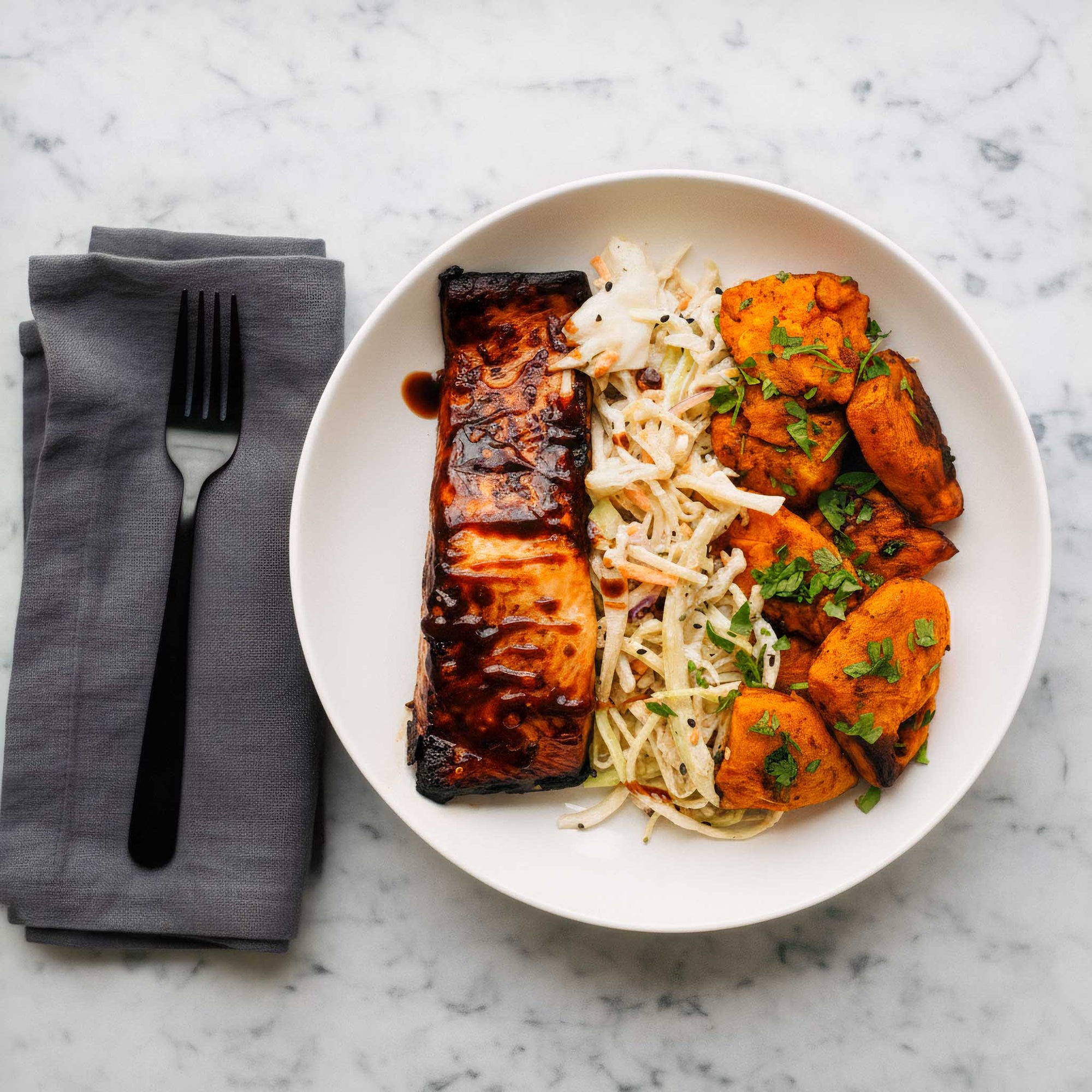 White plate of air fried teriyaki salmon with hot shake sweet potatoes and maple mustard slaw, on a marble surface.