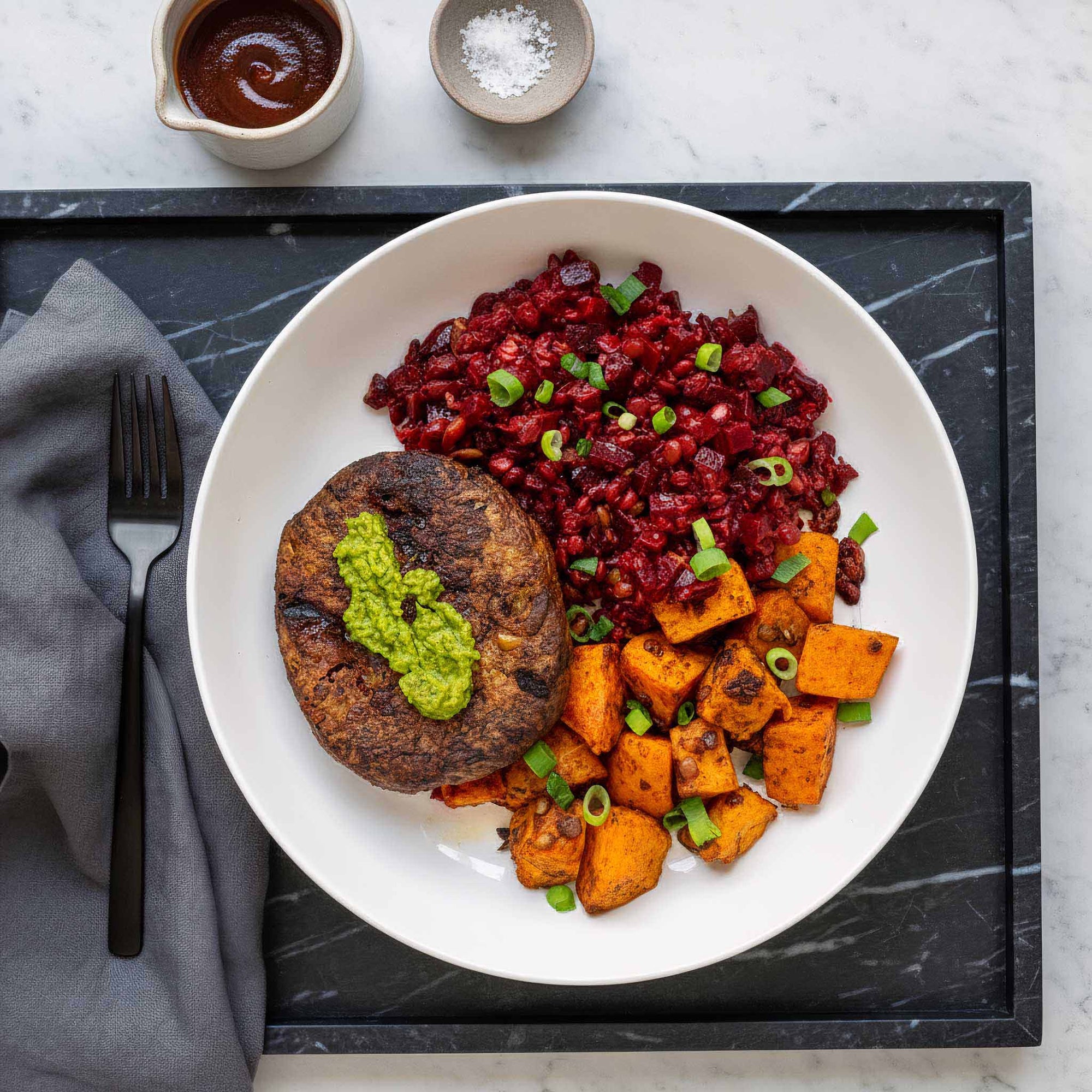 Plated dish with a Soy and cranberry vegan patty, sweet potato cubes, and a beetroot lentil salad on a marble surface. next to a folded napkin and black fork.