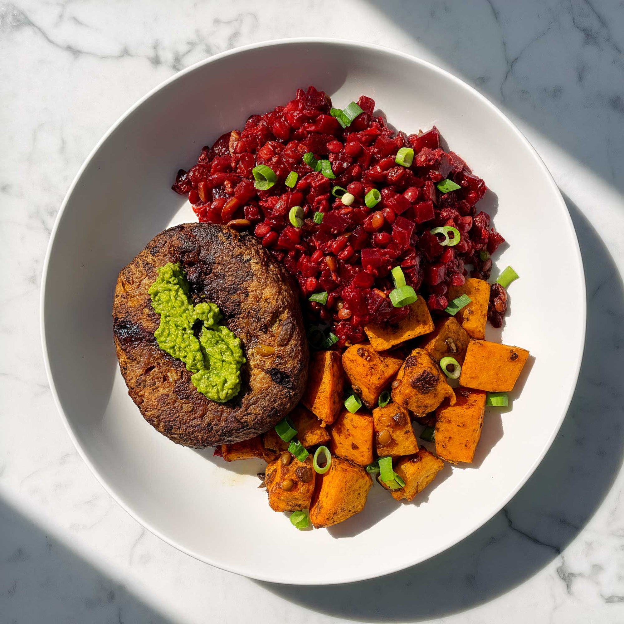 Plated dish with a Soy and cranberry vegan patty, sweet potato cubes, and a beetroot lentil salad on a marble surface.