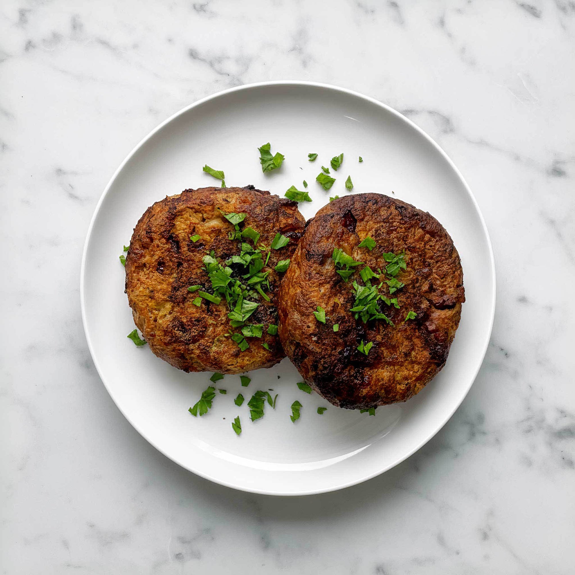 Two browned vegan patties made with soy, caramelised onion and cranberry on a white plate with green herbs on a marble background.