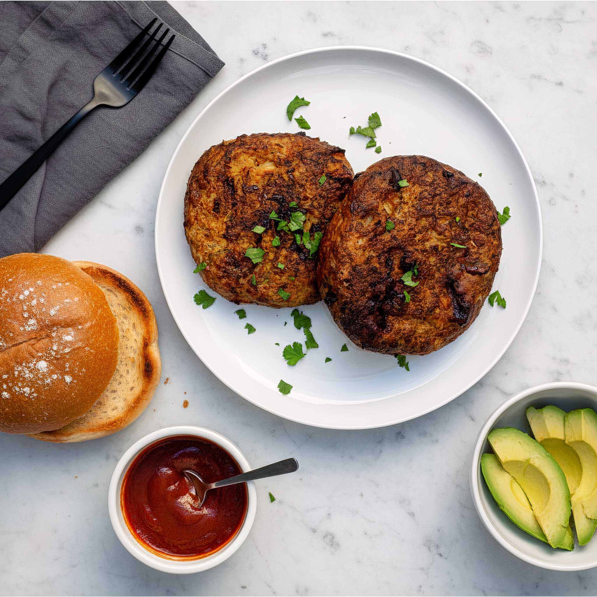 Two vegan soy and cranberry patties on a white plate with a burger bun, ketchup, and avocado slices on a marble surface.